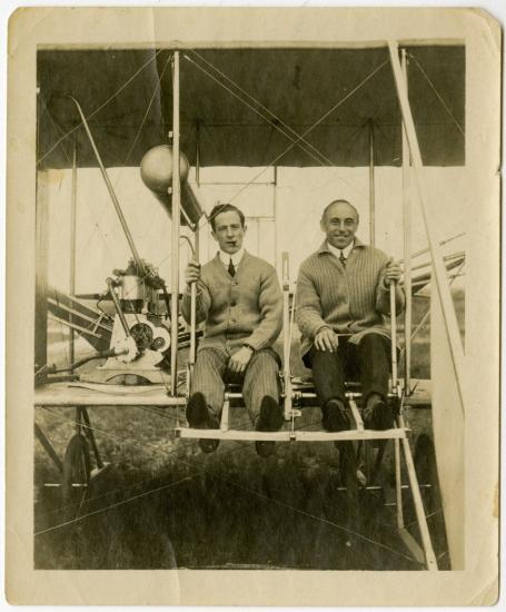 Harry Bingham Brown, a white male pilot, sits in his Wright biplane on the left of another male. Brown is smoking a cigar.