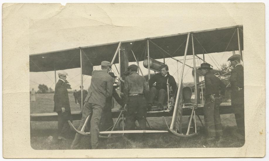 Harry Bingham Brown, a white male pilot, prepares for takeoff in his Wright biplane with the help of several other men.