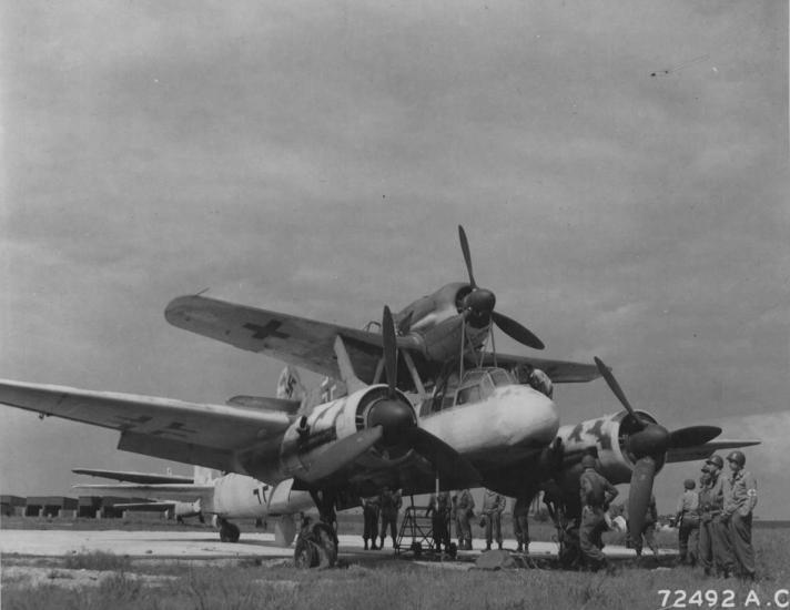 Men stand around a aircraft with two engines which is holding a smaller monoplane with one engine on top of the fuselage.