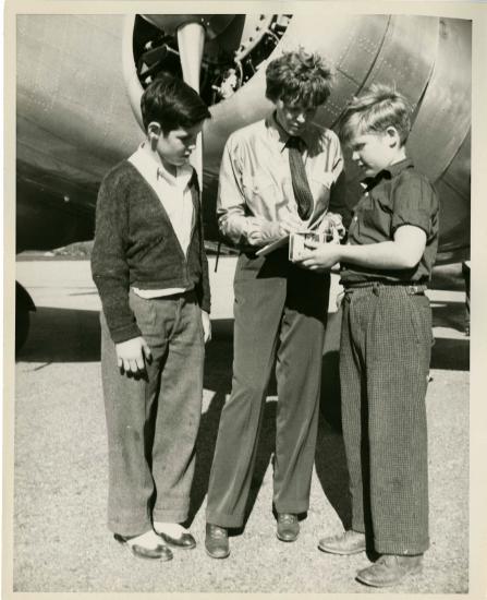 Amelia Earhart, a white woman, stands in between two white people in front of an aircraft. She is signing an autograph for the person on her left (our right).