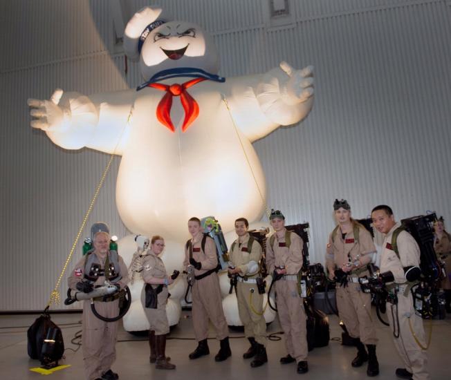 People dressed up in Ghostbusters costumes stand together in front of a Ghostbusters ghost float inside the Museum.