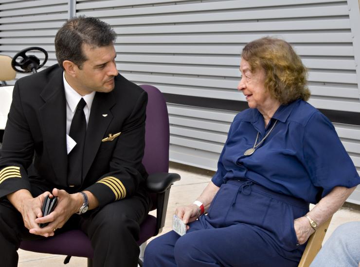 Jerrie Mock, a white female pilot, sits next to and speaks to a male pilot at a Museum event.