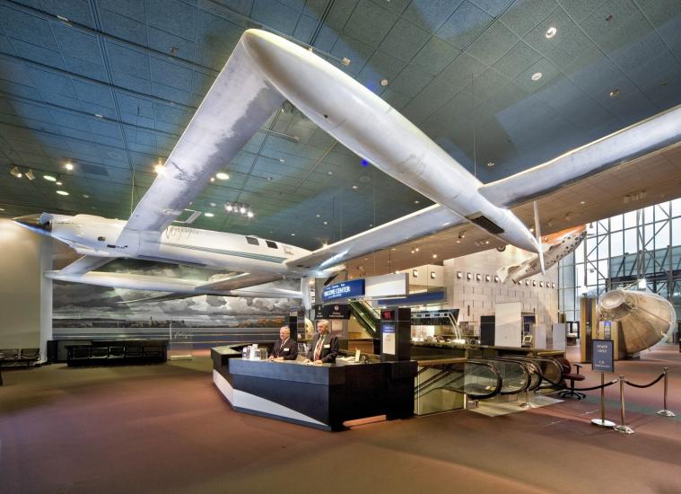 Two docents stand at the museum's welcome desk. A white, twin-boom aircraft hangs above the desk.