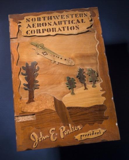 Scrapbook cover featuring a depiction of an aircraft in a downwards position moving toward a desert landscape. The name of the corporation is above the figure, and the president of the corporation who owned the scrapbook has his name written below the figure.