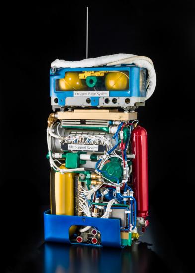 Cutaway view of life support system, a large set of wires and tubes attached to multiple cylinders, and oxygen purge system, which features multiple smaller cylinders around a green tube.