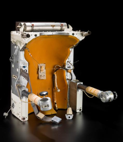 Orange, white, and silver-colored object with silver-colored straps to restrain a human. Arms with buttons at the end of the arm are used to navigate astronauts in space.