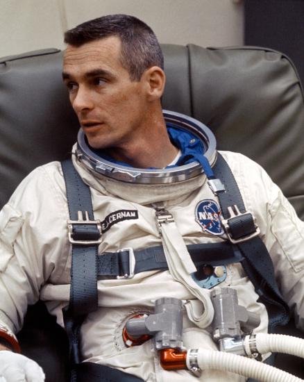 Astronaut Eugene Cernan, a white male, sits while wearing his astronaut suit without the helmet. Multiple life support tubes are seen attached to the suit.