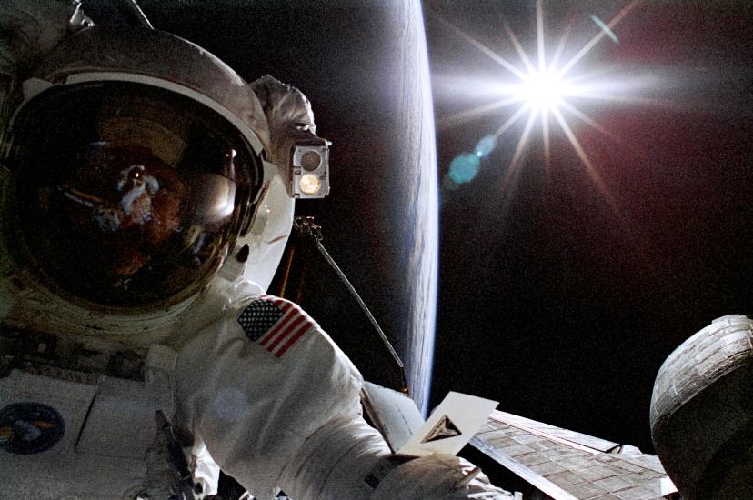 Astronaut Joseph R. Tanner, a white male astronaut, poses during a spacewalk. A silhouette of the Earth can be seen behind him.