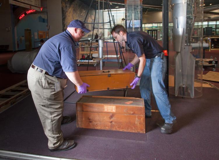 Two Museum staff members work to move a set of wooden boxes out of an exhibit.