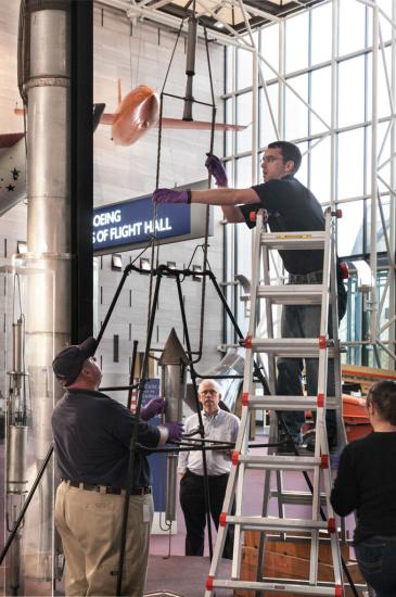 Staff members work to disassemble a primitive rocket from display in the Museum. One person is on a ladder while an other person holds the frame structure together.