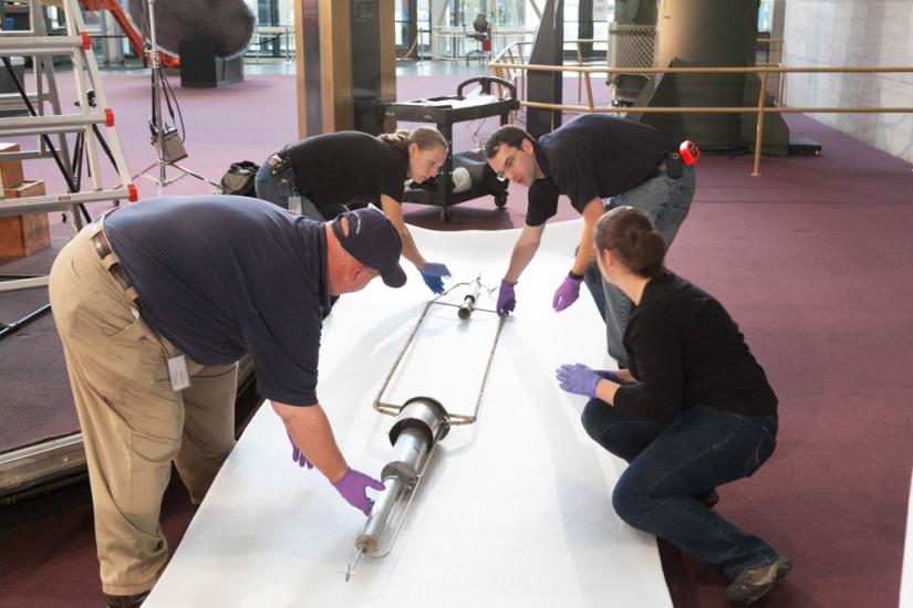 Four Museum staff members work to prepare a primitive rocket for travel away from a museum exhibit.