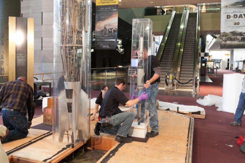 Three Museum staff members work to remove a primitive rocket from a protective case prior to transporting the rocket.