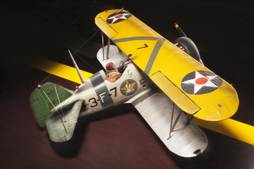 Top sidal view of yellow and silver-colored biplane model aircraft. One white star inside a blue circle is featured on each side of the top wing and identification number "3-F-7" is visible in black paint on the side of the fuselage.