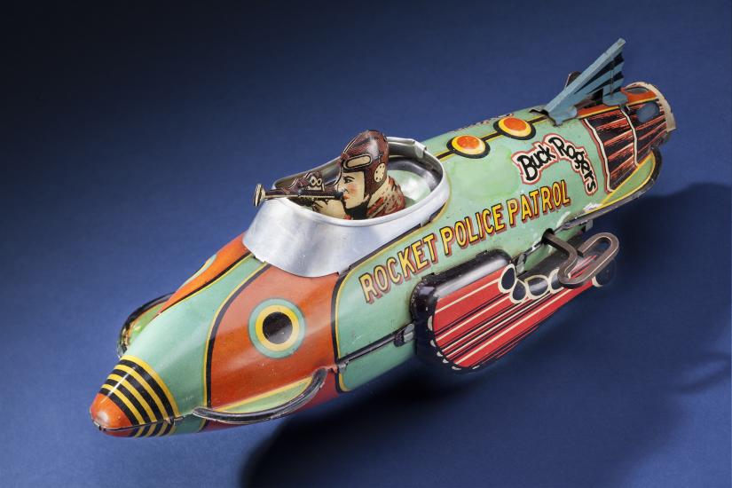 Side view of light green and orange rocket ship figure with character figure in cockpit. "Rocket Police Patrol" is painted on the side of the cockpit in yellow and black.