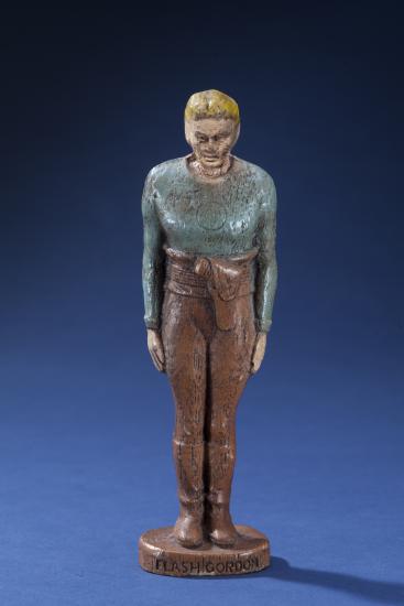 Front view of person-shaped statuette with blue-gray top and brown pants. "Flash Gordon" is painted in black on the front of the base.