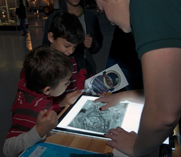 One staff member of the Museum and two younger-aged Museum visitors work together to solve a game. They are looking at an object on a white screen.