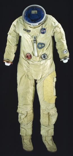 Front view of full-body spacesuit made of tan fabric. Suit does not include gloves or helmet, but it includes the life support tubes. NASA patch on right chest and nametag on left chest.