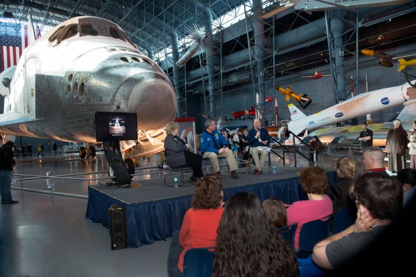 Three Museum staff members host a presentation in front of the Space Shuttle Discovery, a white aircraft designed for space travel, during an open house event.