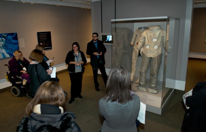 A white female museum conservator speaks to visitors about two tan brown-colored spacesuits on display inside an exhibit about spaceflight.