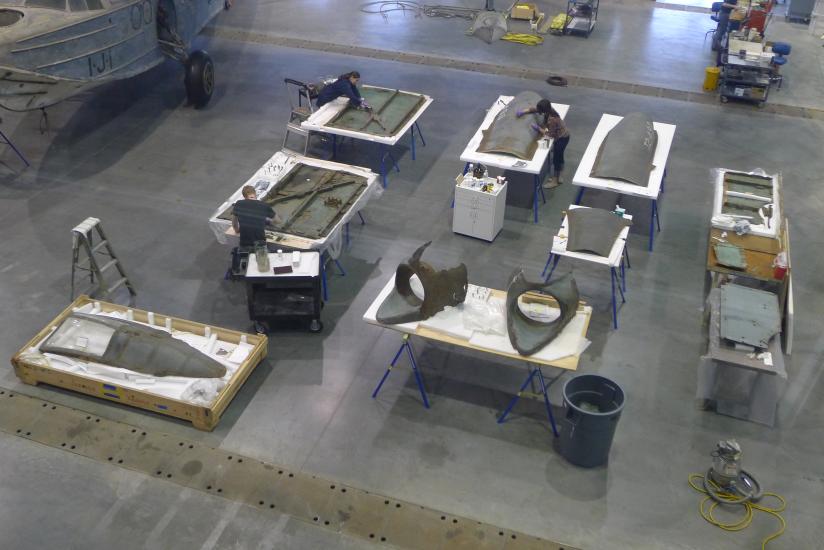 Multiple pieces of a gray, metal aircraft are placed separately on eight tables. Three Museum conservators are performing conservation treatments on separate pieces of the aircraft.