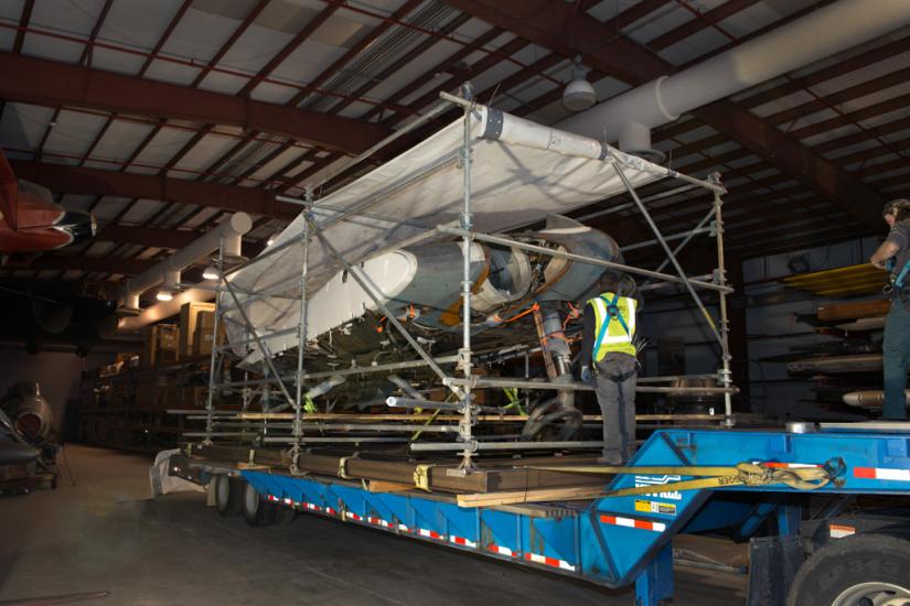 A gray and white metal aircraft, attached to a steel support structure, sits on the long and large bed of a transportation truck. A white tarp protects the top of the aircraft.