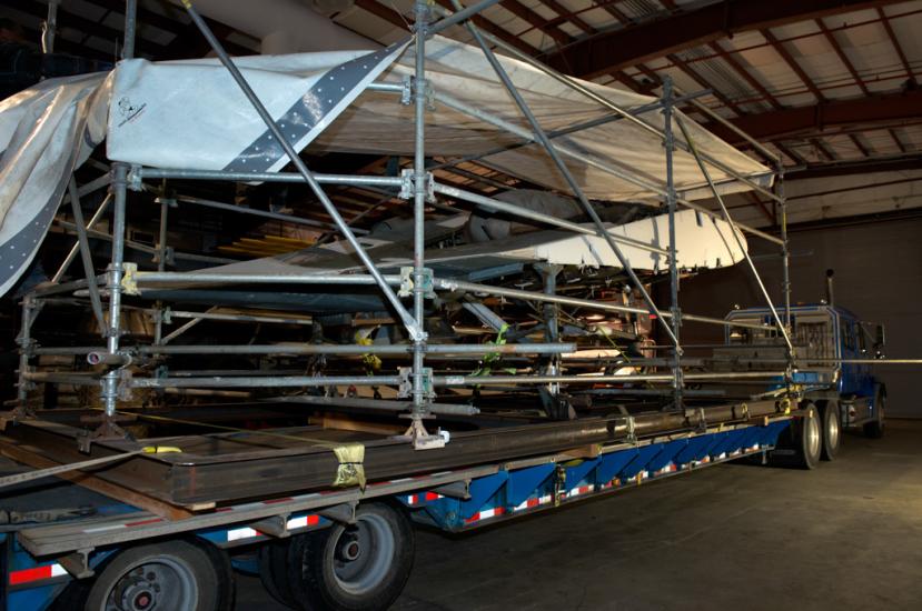 Diagonal back view of the Horton Ho 229 V3, a gray and white metal aircraft, loaded onto a truck for transport. The top of the aircraft is covered by a large white tarp for protection,