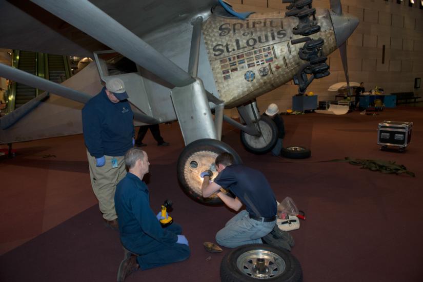 Three Museum staff members work to change tires on the Spirit of St. Louis, a silver monoplane.