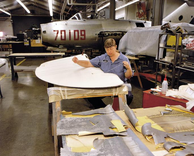 Restoration staff member stitching a control surface is seated holding a line of thread over a beige fabric-covered piece of an aircraft.