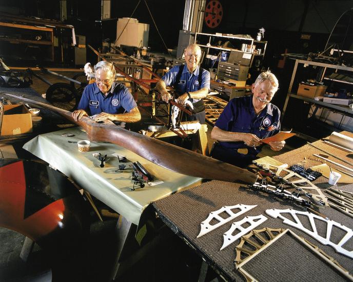 Three restoration volunteers are seated and smiling while working on aircraft parts. A volunteer in the foreground holds sandpaper over the center of a large wood propellor. 