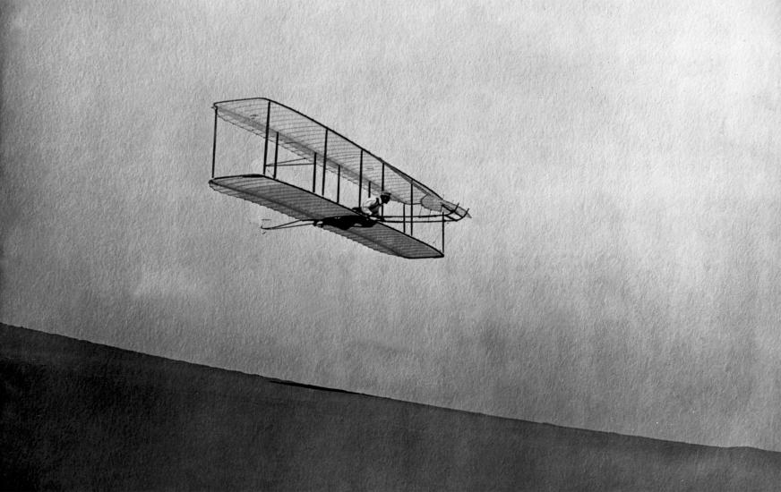 A biplane gliding in the air down a hill. A person lays on top of the bottom wing, perpendicular to it, as they pilot the craft.