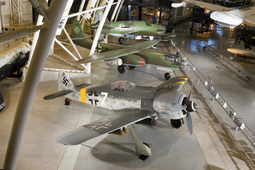 Three German, military aircraft from World War II are displayed next to each other at the Udvar-Hazy Center. The closest aircraft is a silver-colored aircraft and the two aircraft behind it are olive drab green.