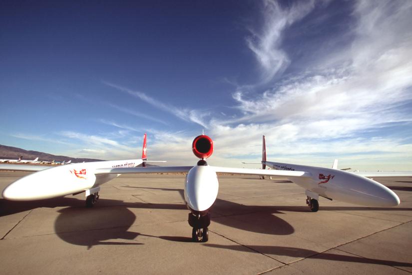 Front view of wide, low winged white aircraft sitting on tarmac with blue sky in the background.