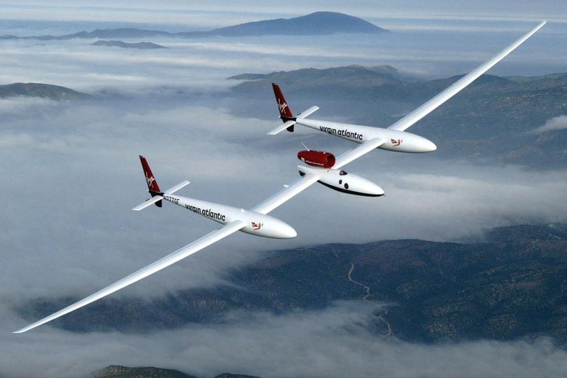 View from above of wide white, low-winged aircraft in flight over cloud-topped mountains.