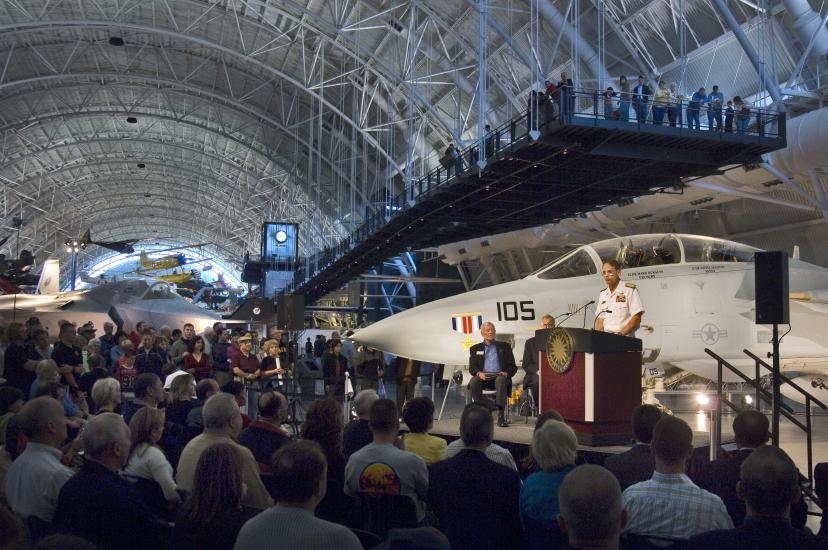 A former director of the National Air and Space Museum speaks in front of a crowd as he welcomes a white fighter jet located behind him at the Udvar-Hazy Center.