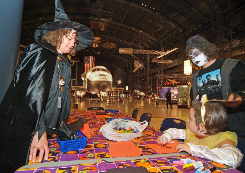 Trick-or-treater vistors stand at a Halloween-themed display with a museum curator dressed in a witch costume.