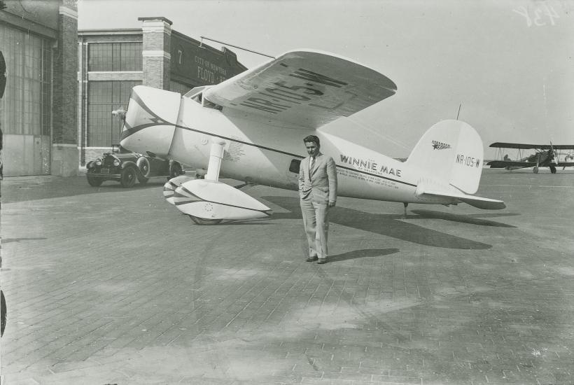 Wiley Post stands on the side of his Winnie Mae aircraft, under its left wing. 