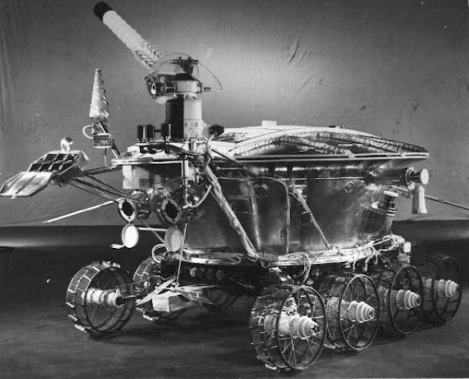 Black and white image of a robot lunar rover with a pie-shaped base and multiple probes, cameras, and antennae attached to the base.