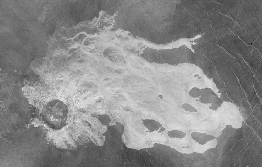 A black and white satellite view of a section of Venus's surface with a large crater impact and lava flows surrounding the crater. The lava flows are lighter in color than the rest of the visible surface.