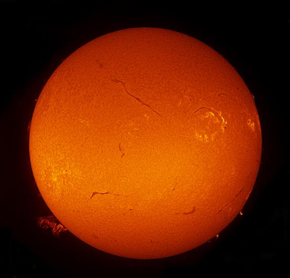 Disk view of the sun with multiple large filaments (dark lines), three sunspot clusters, and one large prominence of plasma rising from the surface visible.