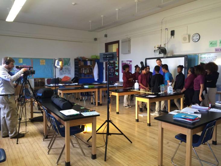 A group of people stand together in a classroom setting on the right while a person films on the left.