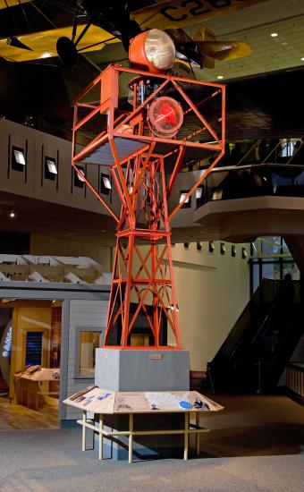 Orange, metal structure holding rotating light used for air travel. Structure is displayed in the Museum.