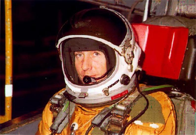 Col. Richard Graham, a white man, sits in an orange and green flight suit and white helmet.