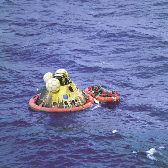 An asymetrical, yellow-colored cylindrical prism sits in a float next to its crew in an orange raft prior to being recovered following the Apollo 11 mission.