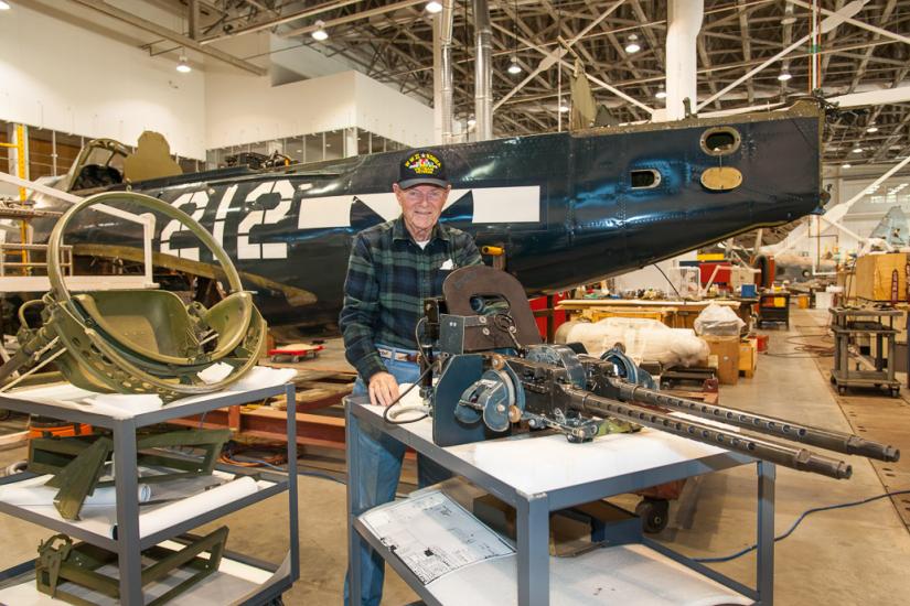 Charles French, a white male veteran, poses in the Restoration Hangar of the museum in front of the Helldiver.