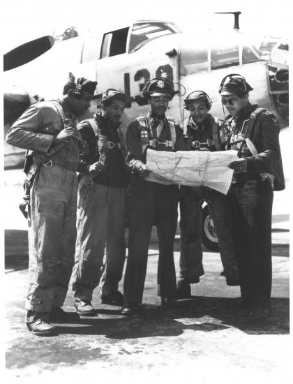 Five Black aviators look at a map. They're dressed in flight gear. There is a plane behind them.