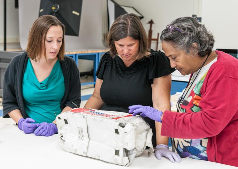 Three female Museum staff members, one of whom is a conservator and the other two are curators, look at a white and red oxygen purge system cover used on the Moon that was recently unsealed.