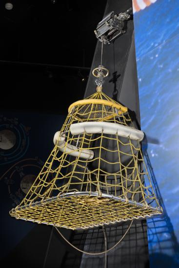 Quadrilateral-shaped metal frame with yellow fabric cords and two white pontoons near top hanging in musuem