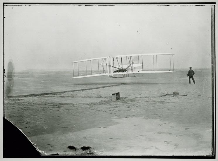 A biplane just barely above the ground with a person standing to the right of it.