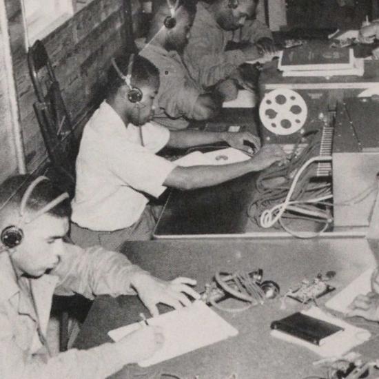 Men sit with headphones on and devices at the center of a table. 