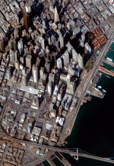 Close-up, satellite view of San Francisco, with high rise buildings to the north and smaller buildings and the San Francisco-Oakland Bay Bridge to the south.